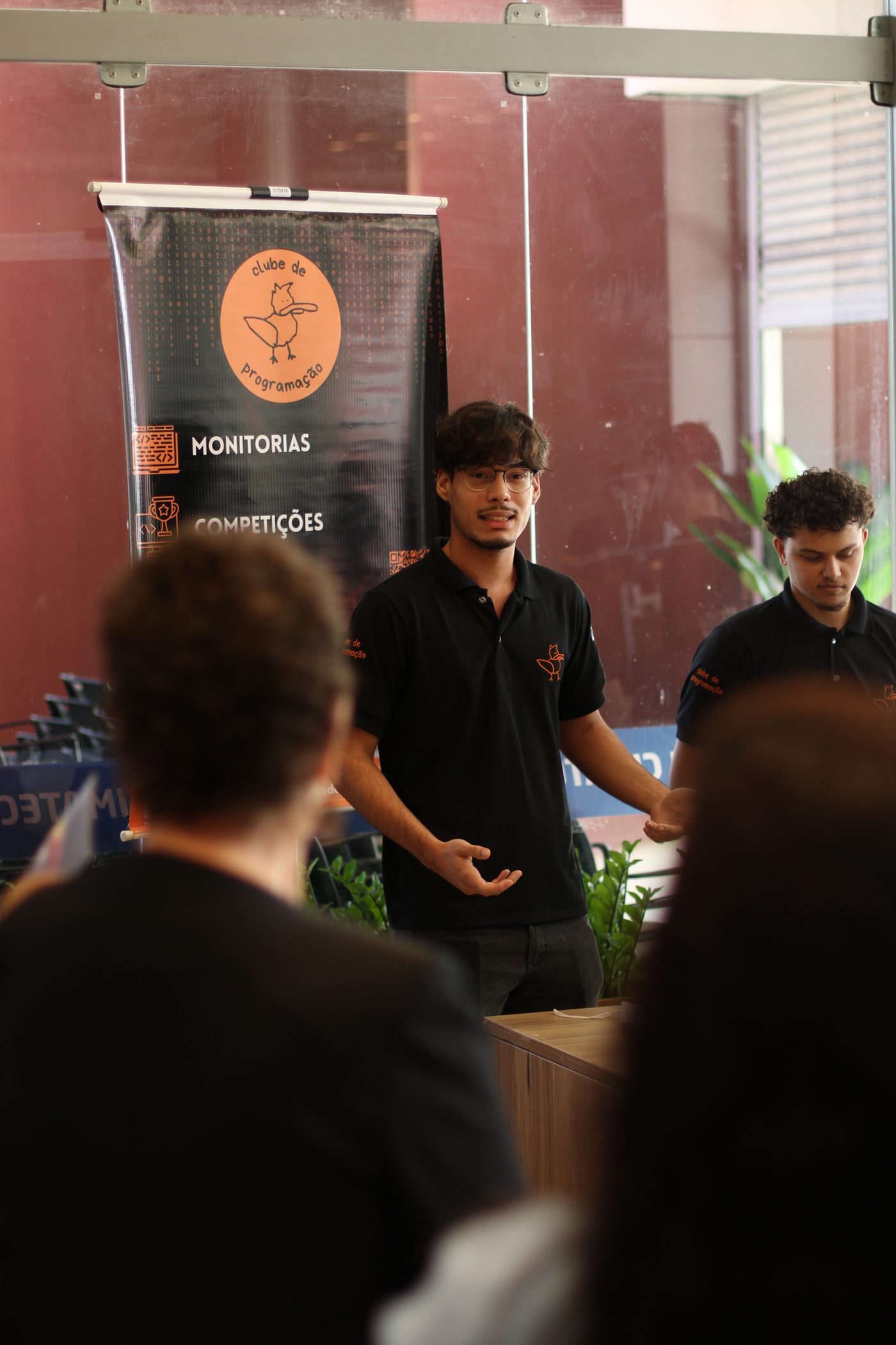 Membro do clube apresentando para a plateia ao lado do banner do Clube de Programação