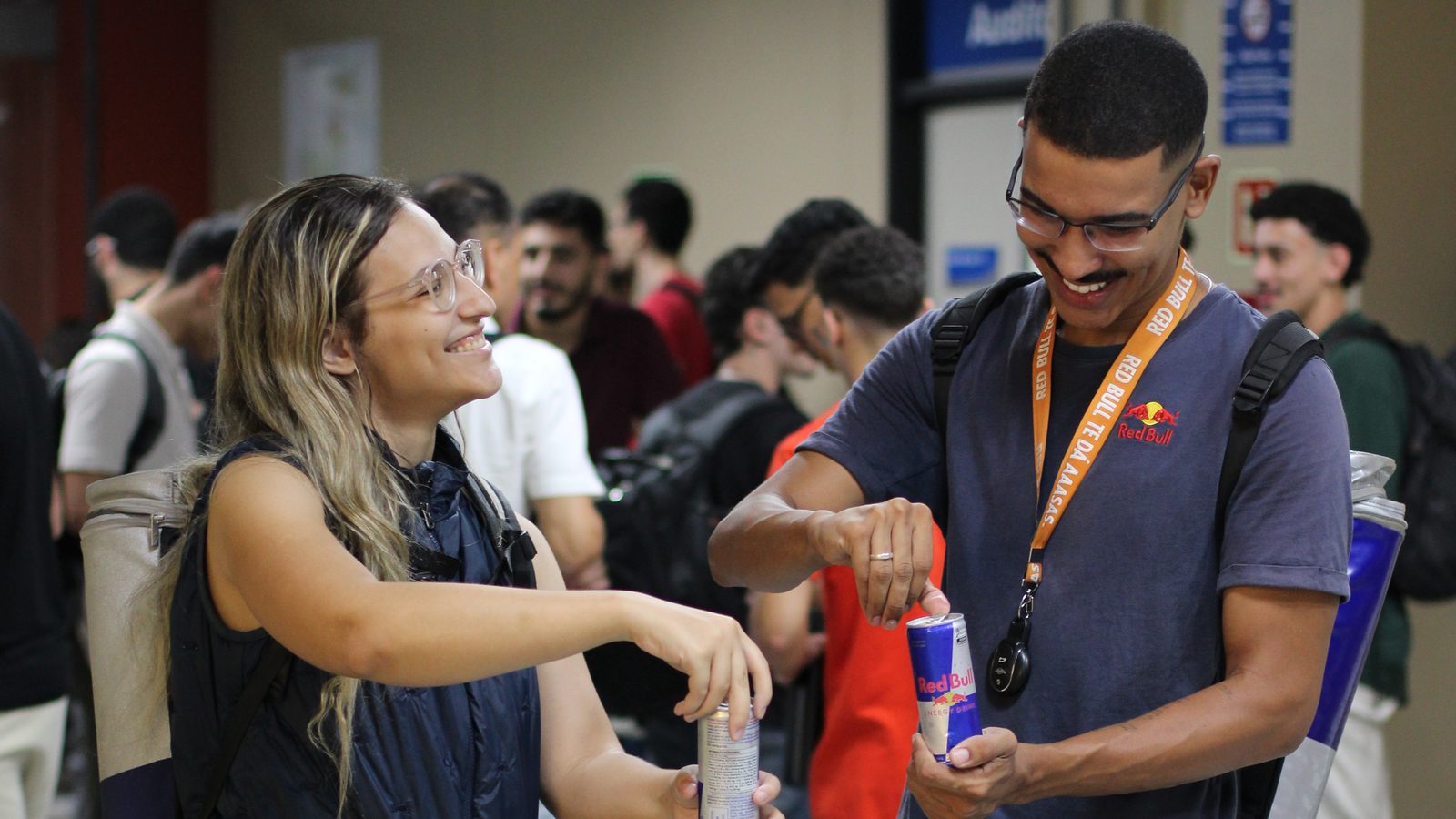 Dois participantes da SEMCOMP conversando e rindo no corredor do evento, com latas distribuídas por um dos parceiros nas mãos